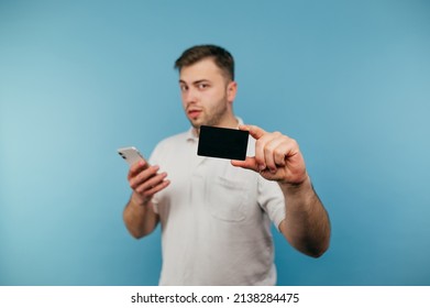 Handsome Man In White T-shirt With Smartphone In Hands Shows Black Bank Card To Camera Isolated On Blue Background