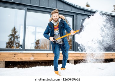 Handsome Man In Winter Clothes Cleaning Snow With A Shovel Near The Modern House In The Mountains