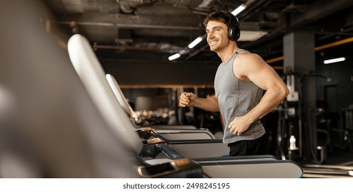 Handsome man in wireless headphones jogging on treadmill at gym and listening music while running, enjoying workout, copy space - Powered by Shutterstock