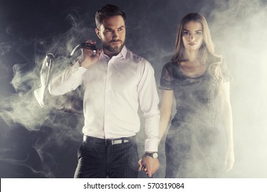 Handsome man and woman in black dress walking through smoke and mist over dark grey background. Studio shot. Toned