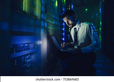 Handsome Man Is Working In Data Centre With Laptop.IT Engineer Specialist In Network Server Room.Running Diagnostics And Maintenance.Technician Examining Server In Big Data Center Full Of Rack Servers