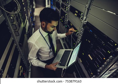 Handsome man is working in data centre with laptop.IT engineer specialist in network server room.Running diagnostics and maintenance.Technician examining server in big data center full of rack servers