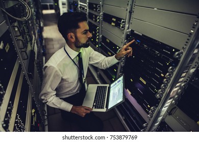 Handsome man is working in data centre with laptop.IT engineer specialist in network server room.Running diagnostics and maintenance.Technician examining server in big data center full of rack servers