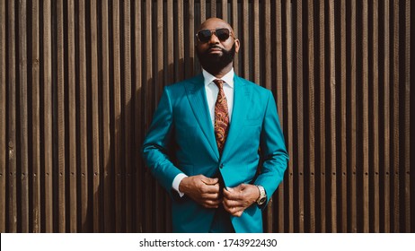 A handsome mature bald bearded African man in a sunglasses and a fashionable blue or teal costume with a tie is standing in front of a wall made of striped wooden timbers and fastening a suit button - Powered by Shutterstock