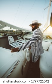 A Handsome Mature Bearded Black Guy In A Summer Hat And A Striped Fancy Costume Is Driving His Luxury Sailboat, Turns The Ship's Wheel With One Hand, And With The Other Hand Takes A Glass Of Champagne