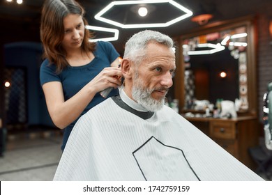 Handsome mature bearded man sitting in barbershop chair while female barber working with hair clipper, making haircut. Barbershop. Beauty salon. Barber tools. Mens haircut - Powered by Shutterstock