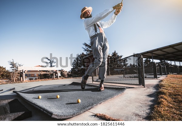 A handsome mature bearded retrosexual black guy in\
trousers with suspenders, white shirt, and hat is got the stance\
and hit the ball with a golf club on a golf field; several yellow\
balls near him