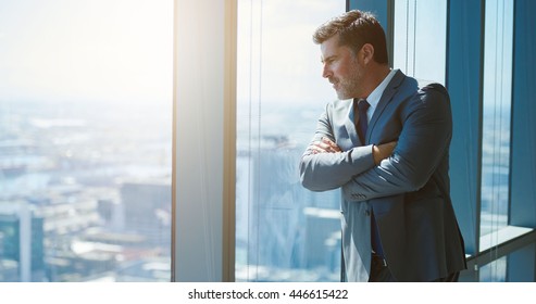 Handsome Mature Business Executive With Attractive Designer Stubble, Standing With His Arms Crossed And Leaning Against A Large Window In A Top Floor Office, Looking Down At City