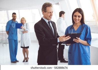 Handsome mature businessman and beautiful young doctor are discussing documents and smiling while standing in the hospital hall - Powered by Shutterstock