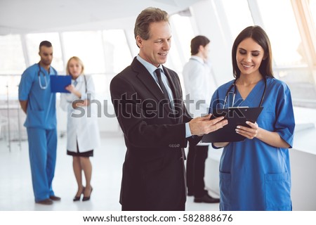 Handsome mature businessman and beautiful young doctor are discussing documents and smiling while standing in the hospital hall
