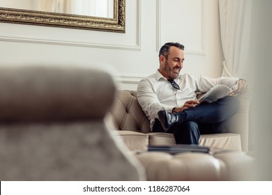 Handsome Mature Businessman Reading A Magazine While Relaxing In Hotel Room. Man On Business Trip Staying In A Hotel.