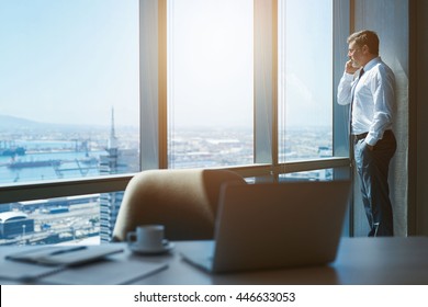 Handsome mature businessman standing next to the large windows of his top floor office, looking at the view of the city while talking on his mobile phone - Powered by Shutterstock