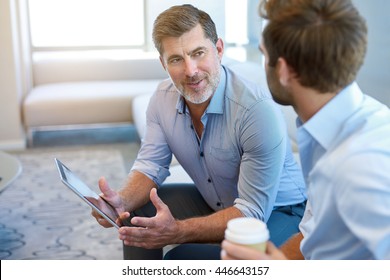 Handsome and mature corporate leader sitting in a business lounge with a young man, offering mentoring advice to him while holding a digital tablet - Powered by Shutterstock