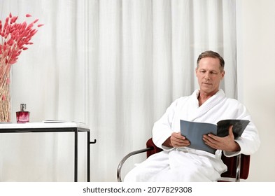 Handsome Mature European Man In White Terry Bathrobe, Sitting On An Armchair And Reading Magazine While Relaxing In Privacy Spa Room Of Wellness Spa Center After Receiving Male Beauty Treatment