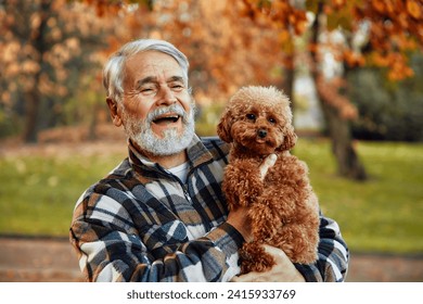 Handsome mature gray-haired man holding dog in his arms in park. Walk with your pet in the fresh air. Grandpa playing with dog. - Powered by Shutterstock