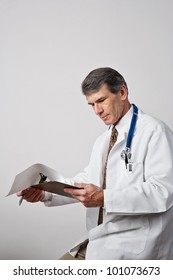 Handsome Mature Male Doctor Reviewing His Notes With Clipboard And Pen. Plain Gray Background.