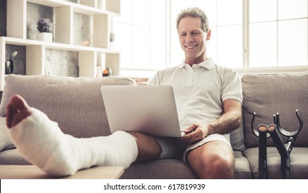 Handsome Mature Man With Broken Leg In Gypsum Is Using A Laptop And Smiling While Sitting On Sofa At Home