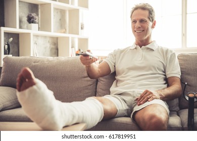 Handsome Mature Man With Broken Leg In Gypsum Is Using A Remote Control And Smiling While Watching TV On Sofa At Home