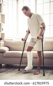 Handsome Mature Man With Broken Leg In Gypsum Is Trying To Move Using A Pair Of Crutches At Home