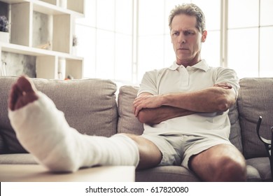 Handsome Mature Man With Broken Leg In Gypsum Is Sitting Seriously With Crossed Arms On Sofa At Home