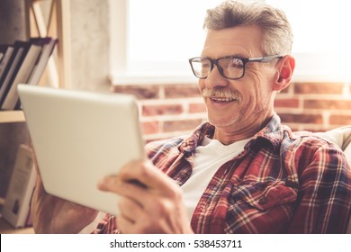 Handsome mature man in casual clothes and glasses is using a digital tablet and smiling while resting at home - Powered by Shutterstock