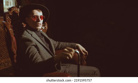 Handsome Mature Man In Elegant Expensive Clothes And Glasses Sits In The Armchair In A Luxury Vintage Apartment. Gangster Man, Mafia. 