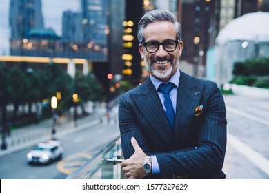 Handsome mature man in glasses and business suit with arms crossed smiling and standing on background of evening New York street while looking at camera