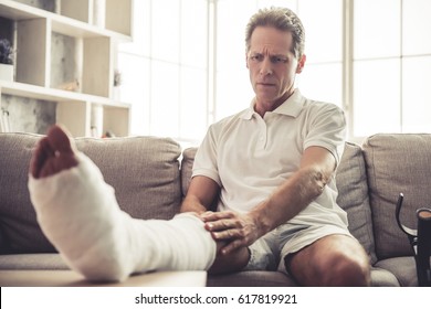 Handsome Mature Man Is Touching His Broken Leg In Gypsum While Sitting On Sofa At Home