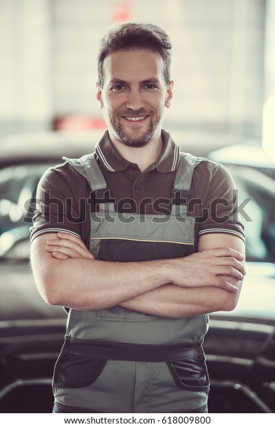 Handsome mechanic in\
uniform is looking at camera and smiling while standing with folded\
arms in auto service