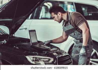 Handsome mechanic in uniform is using a laptop while repairing car in auto service