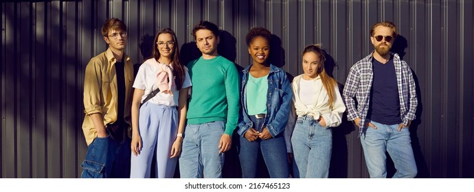 Handsome men and beautiful women in modern comfortable everyday clothes. Group portrait of six diverse relaxed young people in shirts, tees and jumpers leaning on street wall. Casual fashion concept - Shutterstock ID 2167465123