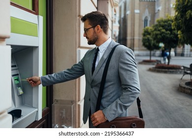 Handsome middle age businessman with eyeglasses standing on city street and using ATM machine to withdraw money from credit or debit card. 
