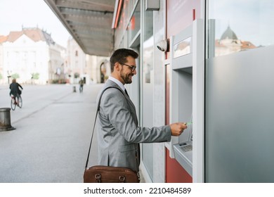 Handsome middle age businessman with eyeglasses standing on city street and using ATM machine to withdraw money from credit or debit card.