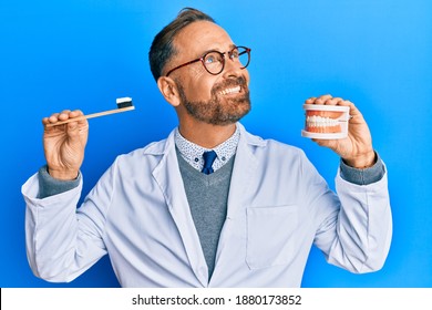 Handsome middle age dentist man holding denture and toothbrush smiling looking to the side and staring away thinking. 