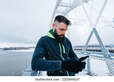 Handsome middle age man with a beard running and exercising outside on extremely cold and snowy day. Sport and fitness motivation theme. He using smart phone to track his activity data. - Shutterstock ID 2203696537