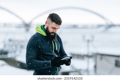 Handsome middle age man with a beard running and exercising outside on extremely cold and snowy day. Sport and fitness motivation theme. He using smart phone to track his activity data. - Shutterstock ID 2319142133