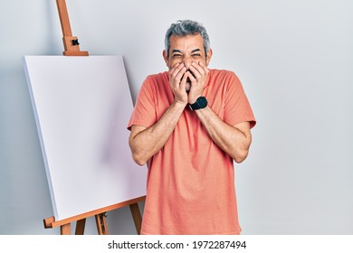 Handsome middle age man and grey hair standing by painter easel stand laughing   embarrassed giggle covering mouth and hands  gossip   scandal concept 