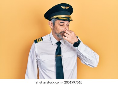 Handsome Middle Age Man With Grey Hair Wearing Airplane Pilot Uniform Smelling Something Stinky And Disgusting, Intolerable Smell, Holding Breath With Fingers On Nose. Bad Smell 