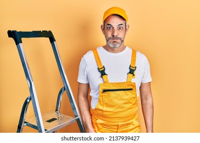 Handsome middle age man with grey hair holding ladder puffing cheeks with funny face. mouth inflated with air, crazy expression. 
