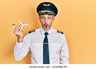 Handsome middle age man with grey hair wearing airplane pilot uniform holding toy plane scared and amazed with open mouth for surprise, disbelief face 