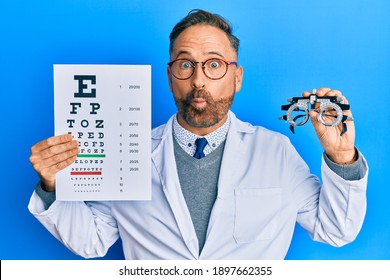 Handsome Middle Age Man Holding Optometry Glasses And Medical Exam Making Fish Face With Mouth And Squinting Eyes, Crazy And Comical. 