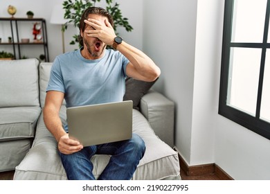 Handsome Middle Age Man Using Computer Laptop On The Sofa Peeking In Shock Covering Face And Eyes With Hand, Looking Through Fingers With Embarrassed Expression. 