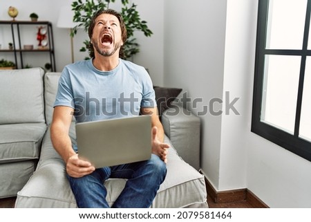 Handsome middle age man using computer laptop on the sofa angry and mad screaming frustrated and furious, shouting with anger. rage and aggressive concept. 