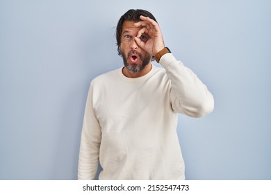 Handsome middle age man wearing casual sweater over blue background doing ok gesture shocked with surprised face, eye looking through fingers. unbelieving expression. 