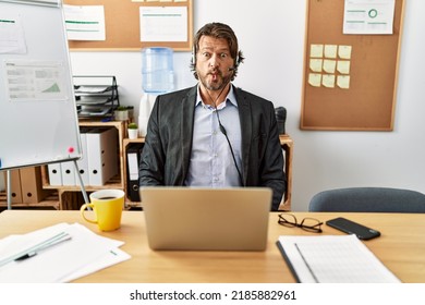 Handsome middle age man wearing call center agent headset at the office making fish face with lips, crazy and comical gesture. funny expression. 
