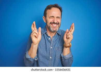 Handsome Middle Age Senior Man With Grey Hair Over Isolated Blue Background Gesturing Finger Crossed Smiling With Hope And Eyes Closed. Luck And Superstitious Concept.