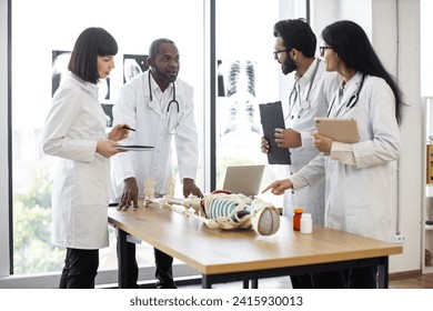 Handsome middle aged African American doctor, anatomy teacher, explaining bones anatomy using skeleton model for diverse students in white coats. Medicine, education, anatomy concept. - Powered by Shutterstock