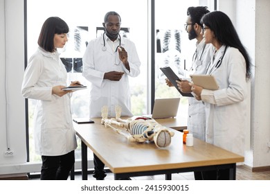 Handsome middle aged African American doctor, anatomy teacher, explaining bones anatomy using skeleton model for diverse students in white coats. Medicine, education, anatomy concept. - Powered by Shutterstock