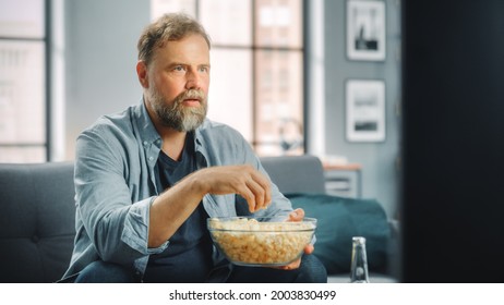 Handsome Middle Aged Bearded Man Sitting On A Couch Watches TV At Home. Focused Fan Eats Snacks, Watches TV Shows, Sports Event, Film, On Demand Streaming Service.