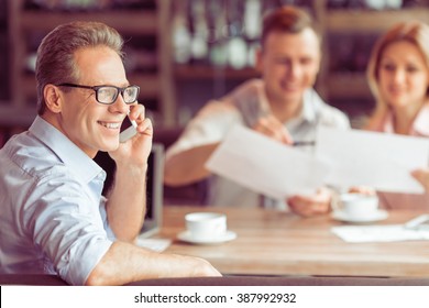 Handsome middle aged businessman in eyeglasses is talking on the mobile phone, in the background young business man and woman working at the restaurant